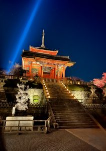 Looking up a flight of stairs at an illuminated red Japanese pagoda at night. A single beacon light shines from the ground behind it up into the sky.