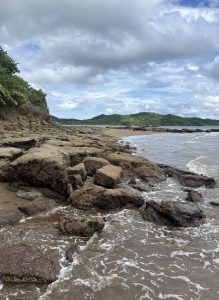 A rocky shoreline with large stones being hit by ocean waves under a cloudy sky, featuring green hills in the background.