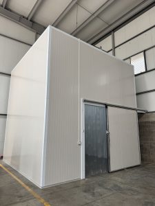 A large, white industrial structure with a flat roof and smooth, panelled exterior is situated within a spacious warehouse. The building features a shiny metallic rolling door on one side and is illuminated by overhead lights.