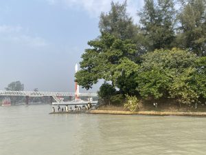 A serene riverside scene with lush green trees on a small island. A group of statues depicting penguin stand near the water's edge, and a red and white tower is visible in the background against a clear blue sky.