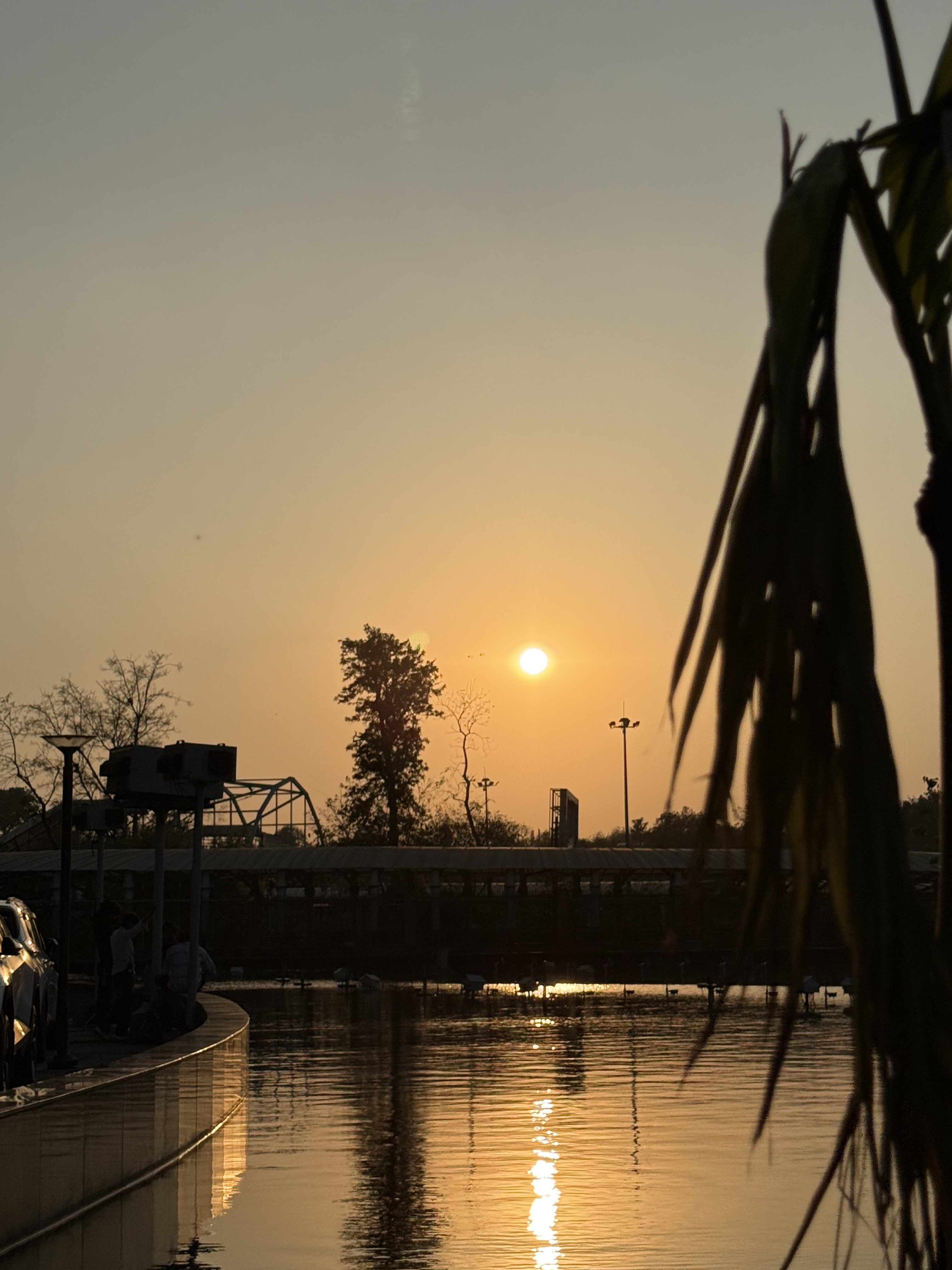 A serene sunset over a calm body of water, with the sun partially obscured by a distant tree on the horizon. The warm orange and yellow hues of the sunset reflect beautifully on the water's surface