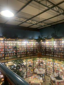 The image depicts an indoor bookstore with tall bookshelves filled with colorful books. The shelves are arranged in a circular pattern, creating a cozy atmosphere, illuminated by hanging lights. There are green plants strategically placed among the bookshelves, adding a touch of nature to the space. The ceiling has a metallic structure, enhancing the modern feel of the bookstore.