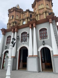 The ornate facade of a colonial-style church with white columns, arched doorways, brick towers, and a clock