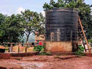 A large black water tank sits on a muddy ground surrounded by lush green trees.