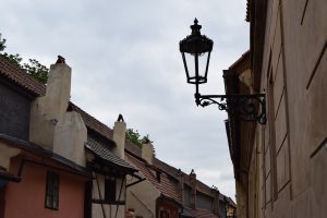 A close-up of an antique black gas lamp in Golden Lane, Prague, with colorful historic houses in the background.