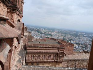 View from a historic Rajasthani fort, showing the intricate red sandstone architecture in the foreground and the sprawling cityscape, featuring many blue-painted houses, stretching out in the background.
