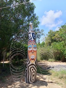 A tall tree trunk is painted with colourful and black-and-white swirling patterns. A wire structure shaped like wings is attached to the sides of the trunk. The sculpture stands on sandy ground in a clearing surrounded by green trees and bushes under a clear sky.