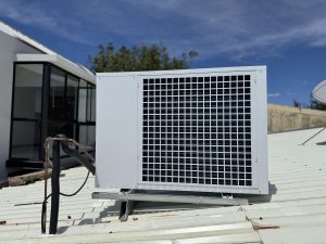 A large air conditioning unit is situated on a sloped metal roof, with a clear blue sky in the background. The unit has a white exterior with a grid pattern on the front, and pipes connected to it.