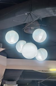 A cluster of white globe-shaped lantern lights hangs from a dark ceiling, providing bright illumination in an indoor space.
