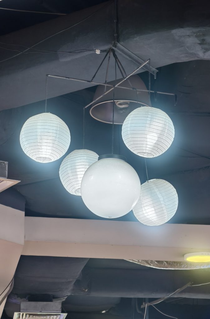 A cluster of white globe-shaped lantern lights hangs from a dark ceiling, providing bright illumination in an indoor space.