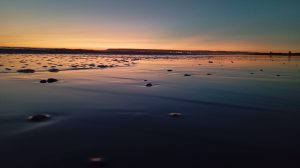 A tranquil sunset over a calm shoreline, with soft orange and blue hues blending across the sky. The shallow water reflects the fading light, scattered with small rocks, while distant land and faint silhouettes line the horizon.