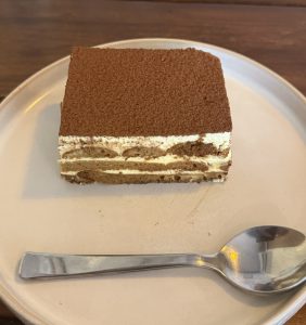 A slice of tiramisu on a beige plate with a metal spoon beside it. The dessert has creamy layers dusted with cocoa, set on a wooden table.