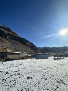 A scenic view of a landscape featuring a patch of snow-covered ground leading to a small, calm lake. In the background, rugged mountains rise under a clear blue sky, with a bright sun shining in the upper right corner. A wooden picnic table is situated near the water's edge, surrounded by sparse vegetation and rocky terrain. Lake Gothard in Switzerland