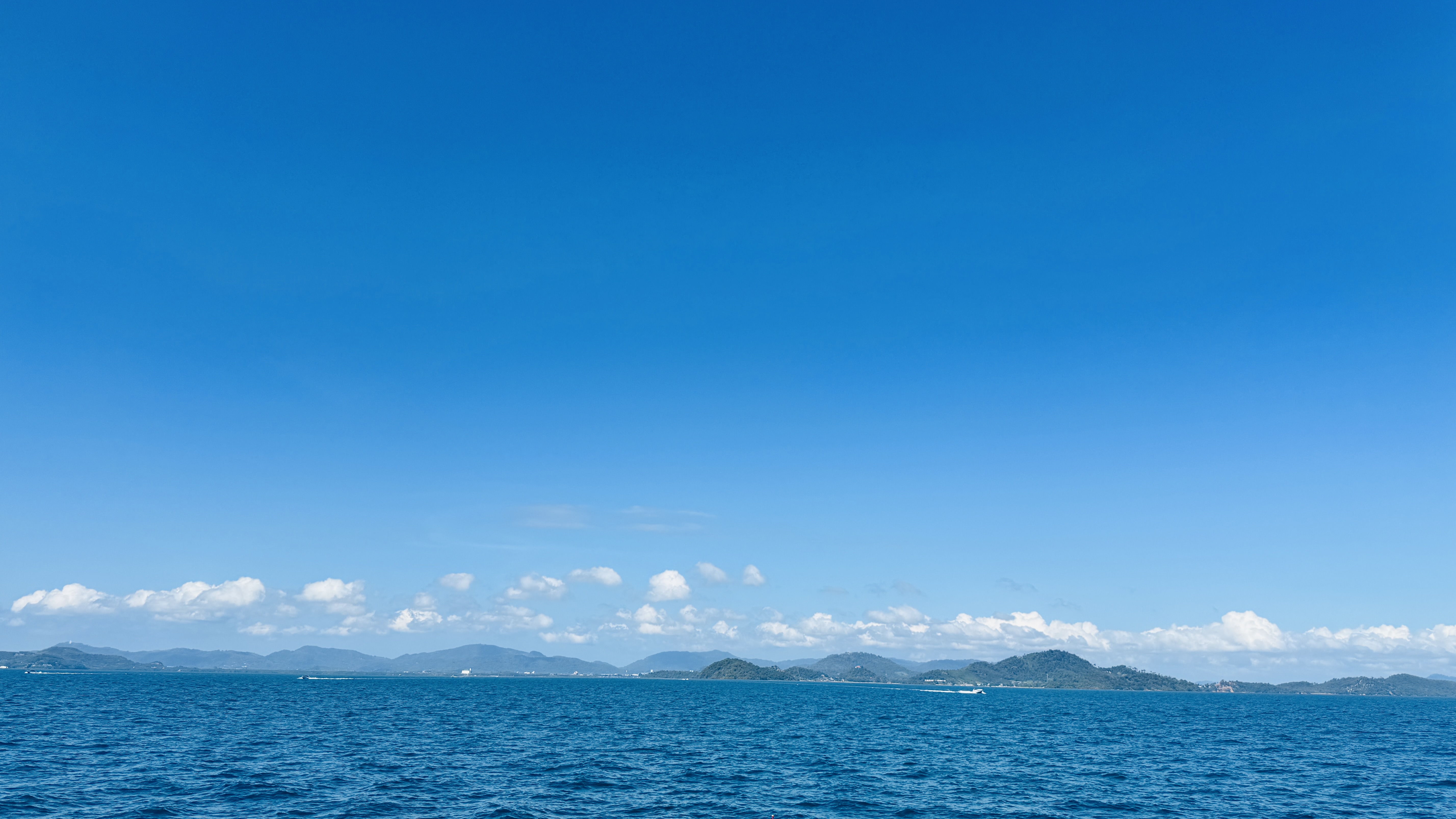 Wide view of a deep blue ocean under a clear sky, with a distant green coastline, small clouds on the horizon, and calm rippling water on a sunny day.