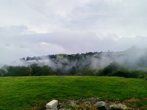 A misty landscape featuring rolling green hills and patches of trees, partially obscured by clouds and fog. In the background, a small cluster of buildings is visible among the greenery. The scene is tranquil, with a soft overcast sky providing muted lighting.
