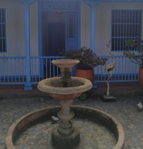 charming courtyard featuring a weathered stone fountain with two basins, surrounded by a cobblestone floor arranged in intricate patterns. Behind the fountain stands a building with cream-colored walls and vivid blue wooden details columns, railings, window frames, and a door evoking a colonial architectural style.
