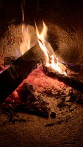 Two thick logs burn in a stone fireplace, their charred surfaces feeding tall orange and white flames. Deep red embers glow beneath, scattered ash and charcoal fragments surrounding them in the darkness.