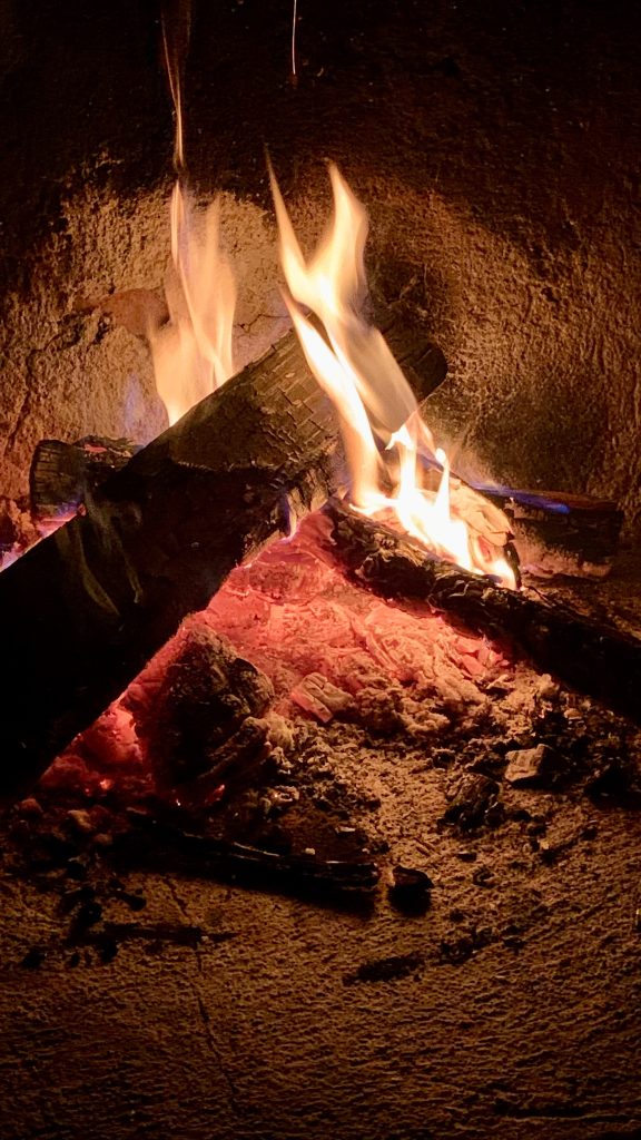 Two thick logs burn in a stone fireplace, their charred surfaces feeding tall orange and white flames. Deep red embers glow beneath, scattered ash and charcoal fragments surrounding them in the darkness.