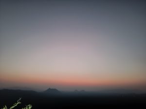 A calm twilight landscape with a blue-to-orange sky, misty distant mountains, faint valley lights, and a peaceful cinematic wide view.
