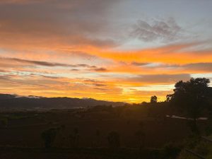A stunning sunset over a rural landscape, featuring a vibrant sky painted with orange, pink, and purple hues. Silhouettes of trees and mountains can be seen in the foreground, while the horizon glows warmly as the sun sets behind distant hills. The fields below are dark brown, hinting at farmland, and clouds add texture to the colorful sky.