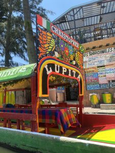 A vibrant, colorful boat decorated with the names "Christopher" and "Lupita" prominently displayed. The boat features intricate designs, including flowers and patterns, with multi-colored tables covered in checkered cloths. In the background, a building can be seen, along with trees and other boats. The overall setting appears to be a lively area near a body of water.