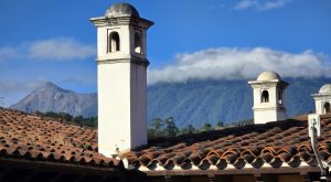 Clay tile roofs and white colonial-style chimneys set against misty mountains and a deep blue sky.