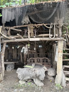 A rustic wooden structure with a layered roof made of foliage and netting. The lower level features a carved wooden bench and table, alongside tree stump stools.