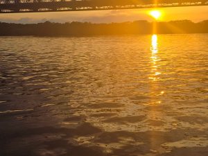 A serene sunset over a body of water, with golden hues reflecting off the rippling surface. In the background, a bridge is silhouetted against the sun as it dips below the horizon, creating a warm and calming atmosphere. The surrounding landscape features dark outlines of trees, contributing to the tranquil scene.
