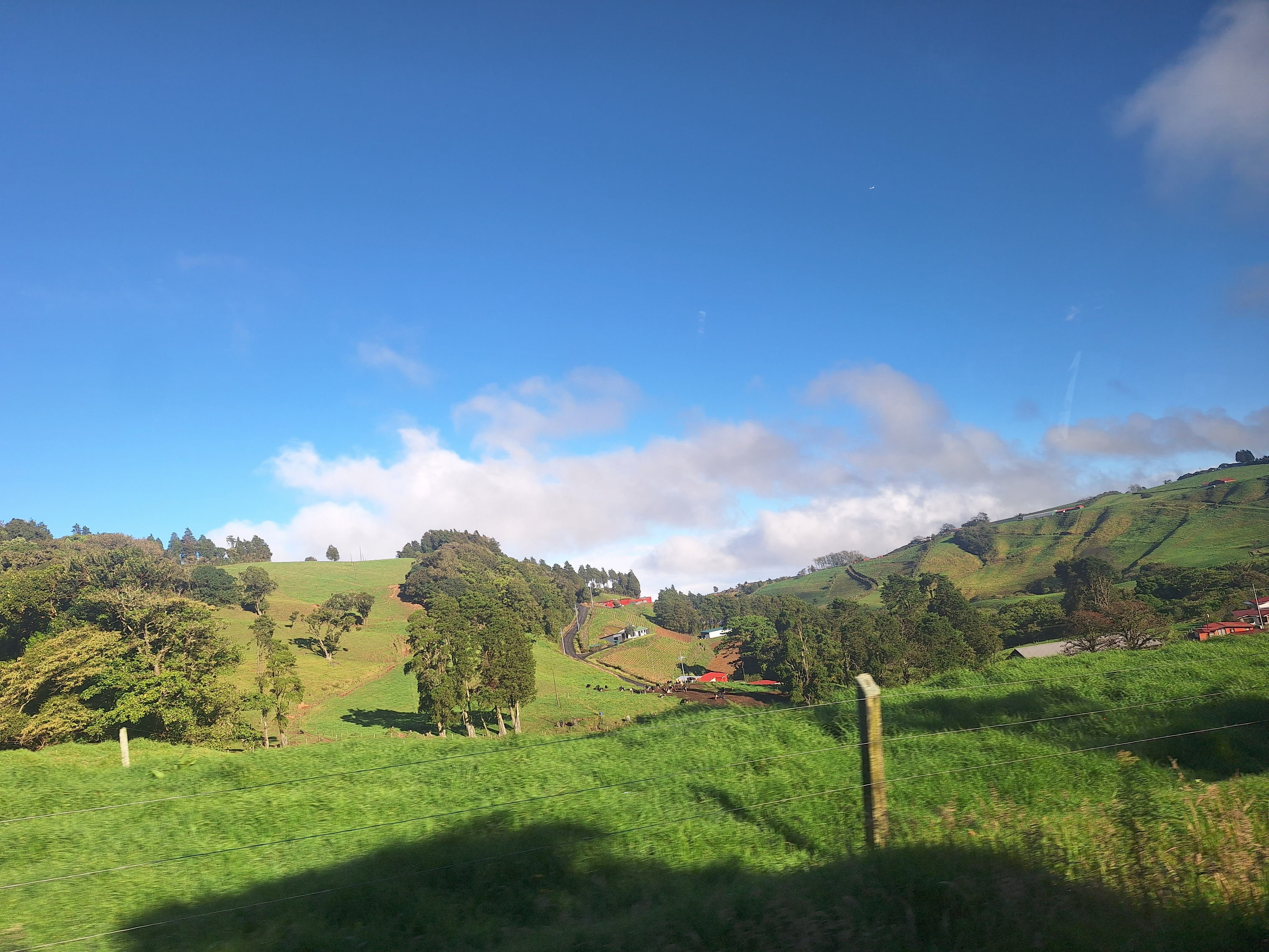A lush, green landscape stretches out under a bright blue sky with a few scattered clouds. Rolling hills are dotted with patches of trees and farmland, while colorful farm buildings are visible in the distance.
