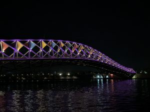 Arched bridge at night, illuminated with purple lights and colorful triangular panels, spanning dark water.