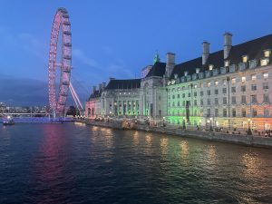 A twilight view of a large, illuminated london eye wheel and a grand historical building along a riverbank, with vibrant red and green lights reflecting on the water.
