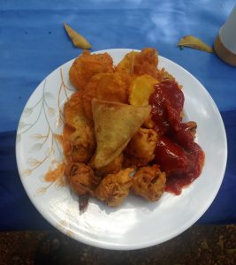 A white leaf-patterned plate with golden fried snacks like bhajias and a samosa, served with tomato ketchup on a blue surface with dry leaves in the background.