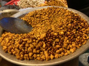 A large metal tray filled with spicy, golden-brown snack mixture including peanuts and crisp bits. A black ladle rests inside the tray, evoking a vibrant market scene.