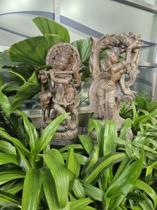 Two detailed wooden sculptures (Lord Krishna and Radha) surrounded by lush green leaves, blending art and nature. A calm, artistic scene captured at Mumbai Airport. 