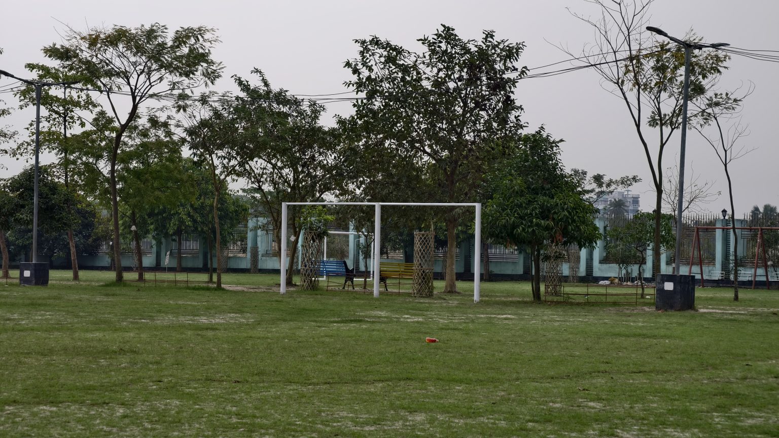 A green park field with several young trees and a white goal post structure under an overcast sky.