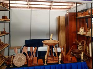 An exhibition booth displaying detailed wooden crafts, including mandala-carved plates, wooden feathers, rolling pins, and key holders, arranged on a blue-skirted table with wooden doors and a dark blue sofa in the background.