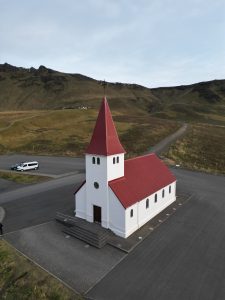 Small church with a red roof in Iceland, surrounded by open landscape, roads, and hills.