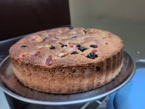 A round cake displayed on a silver plate, featuring a textured, golden-brown exterior. 