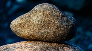A detailed eye-level photo shows a smooth oval granite rock balanced on a larger stone. The rock has a speckled gray, tan, and black texture.