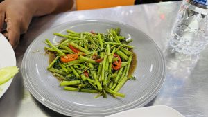 A gray plate of stir-fried morning glory with red chili peppers in brown sauce, served on a metal table with other dishes and a water b