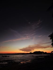 A serene beach scene at sunset, showcasing a vibrant gradient of colors in the sky, transitioning from deep indigo at the top to warm oranges and yellows near the horizon. Silhouetted against the colorful sky are coastal rocks and a small island, with gentle waves lapping at the shore. 