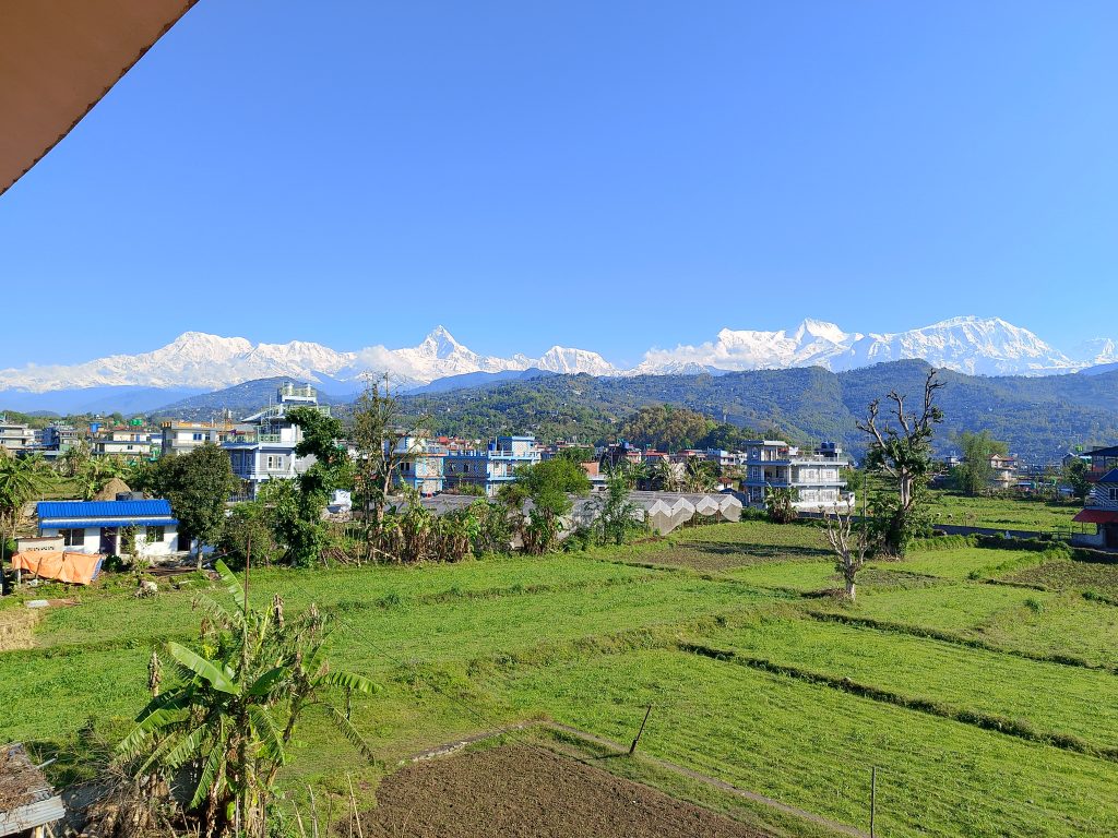 A scenic view of a rural town with green fields in the foreground, scattered houses, and snow-capped mountains in the background under a clear blue sky.
