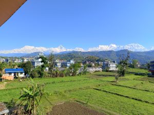 A scenic view of a rural town with green fields in the foreground, scattered houses, and snow-capped mountains in the background under a clear blue sky.