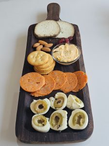 A dark wooden serving platter arranged with crackers, sliced deli meats, almonds, dried cranberries, a small bowl of dip, and herb-crusted cheese.
