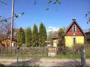 A bright yellow house with a red roof stands behind a wooden fence. Tall green trees line the fence in front of the house, under a clear blue sky.