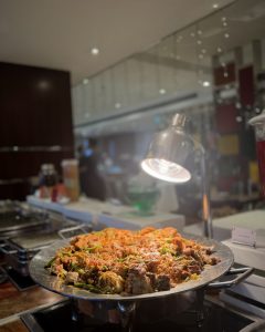 A large, silver serving platter filled with flavorful rice mixed with a variety of vegetables and pieces of meat. The dish is enhanced by a warm, focused light above it, illuminating the vibrant colors.