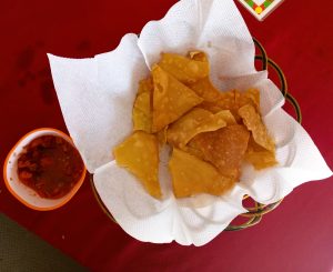 Thin, fried dough triangle nachos are served in a basket with a white napkin, with a small bowl of red tomato salsa on the side.