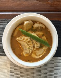 A white bowl filled with spicy orange soup containing dumplings, topped with a green garnish, placed on a black-and-white checkered placemat.