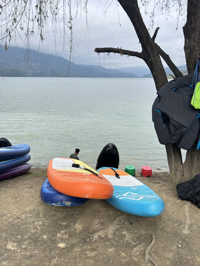 Kayaks are resting beside the lake shore, with a person standing nearby, looking out across the calm water.