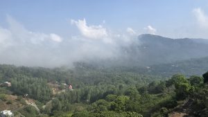 Lush green valley with dense trees and scattered houses, partially covered by white clouds and mist, under a blue sky.
